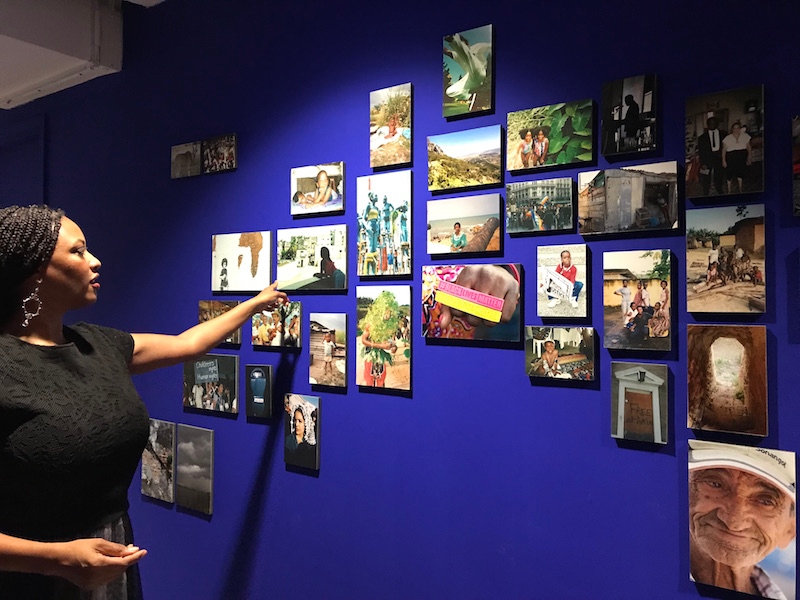 Black woman stands in front of and points to photographs arranges on a blue wall of a gallery